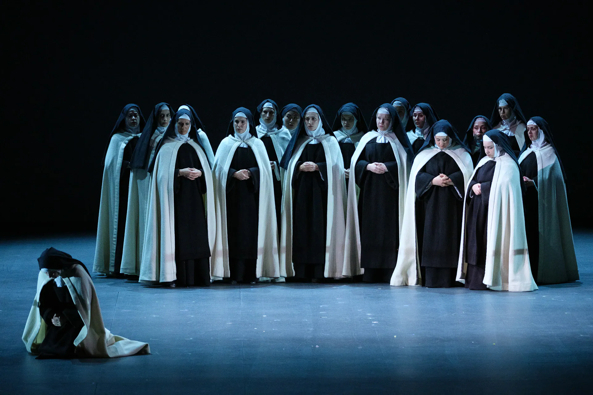 Le carmelitane in fila sotto una luce azzurra, mentre una consorella è inginocchiata in primo piano.