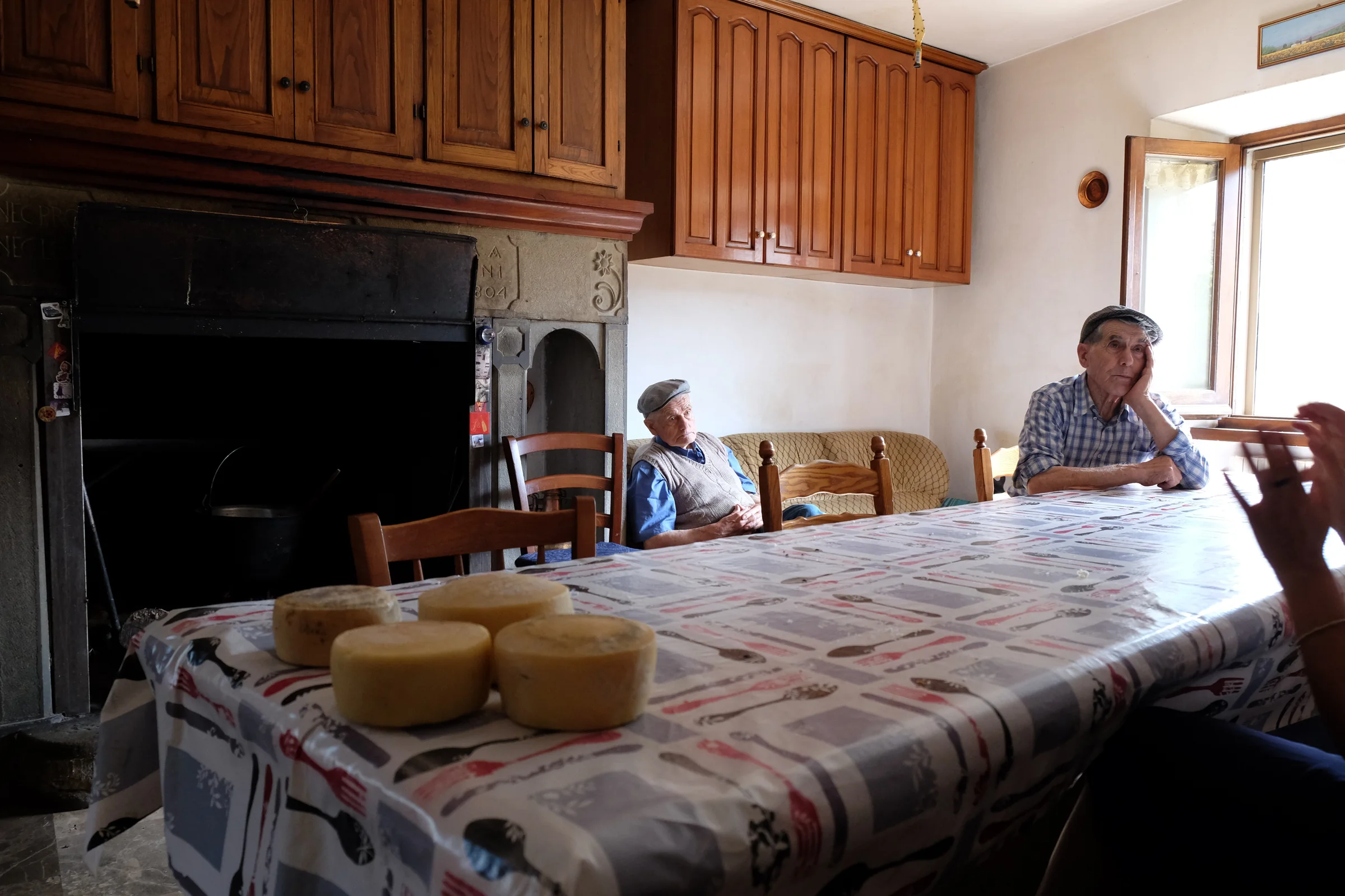 A kitchen with cheese wheels on the table and two men sitting by the fireplace