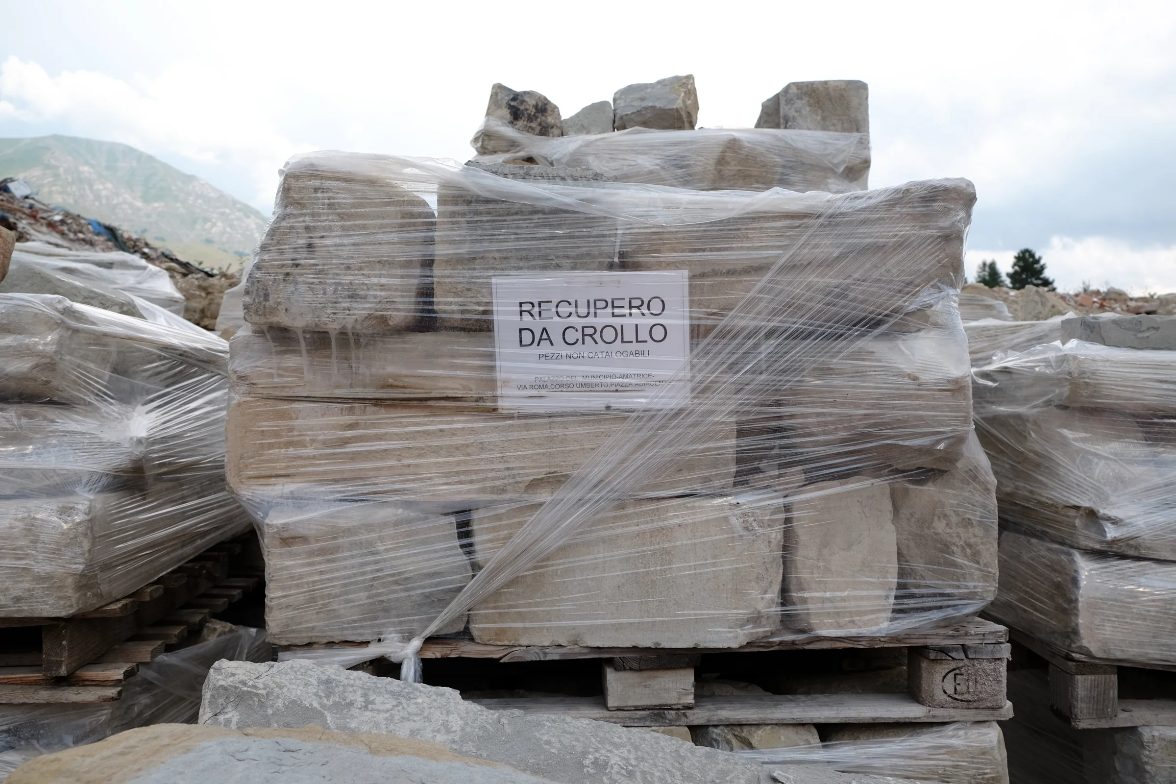 Pallets of stones wrapped in plastic with a sign reading Recupero da crollo