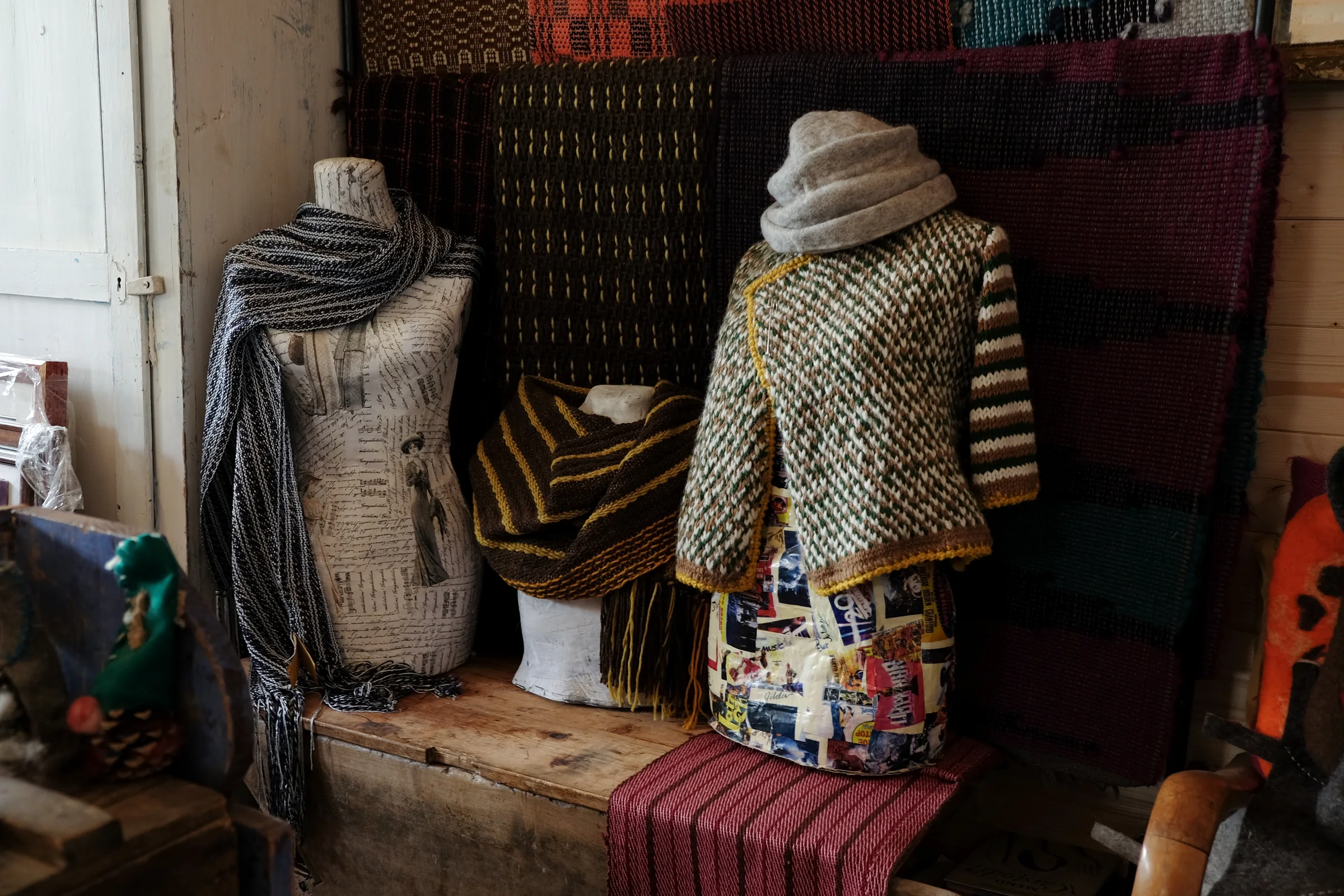 Scarves and a woollen cape on three dress forms, in front of woven cloths