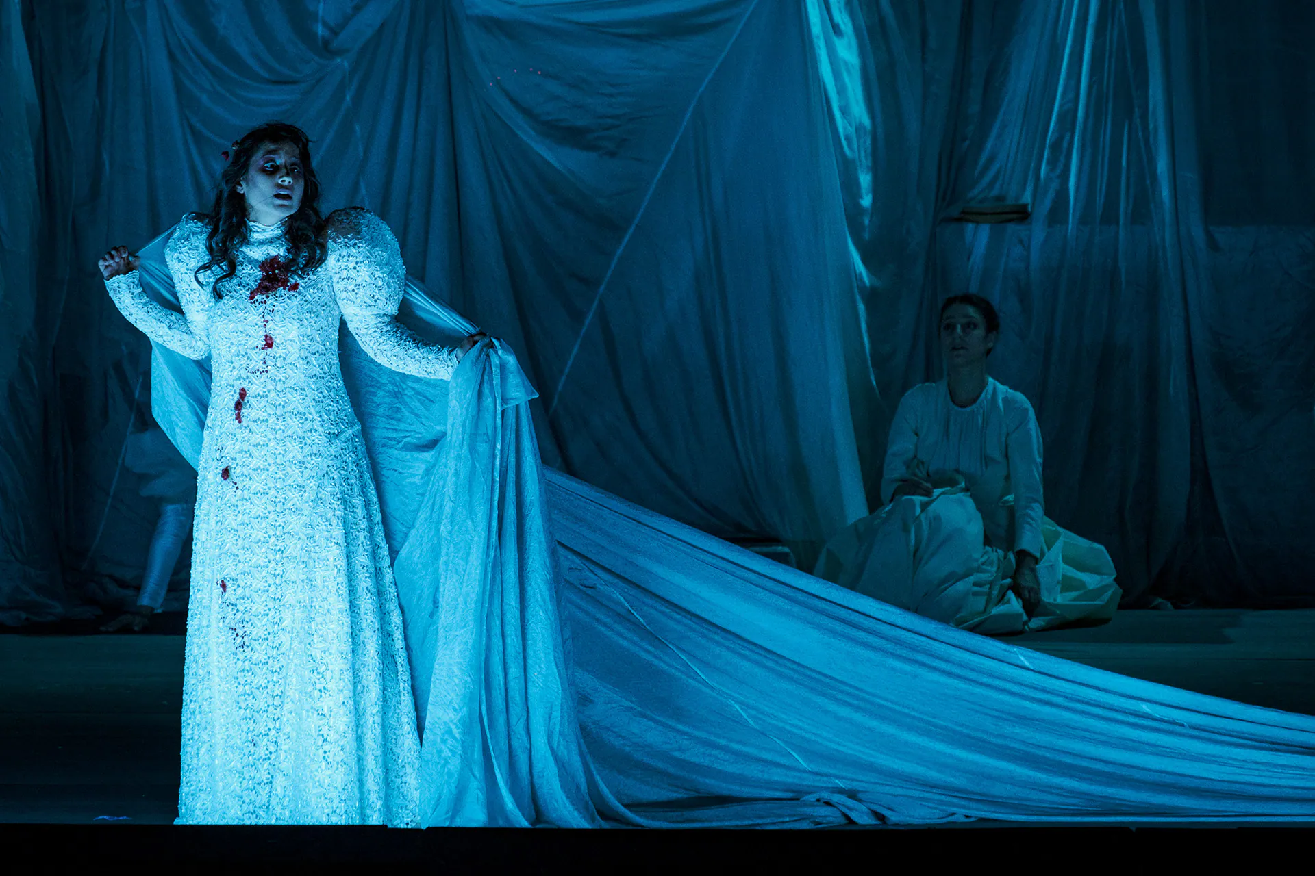 Ophelia in a white gown stained with red under blue light, a seated figure in the background, on stage in Hamlet at Teatro Regio di Torino.