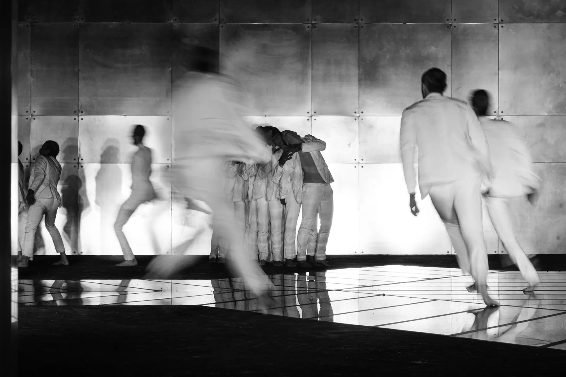 Danzatori vestiti di bianco in movimento sotto luci intense, fotografia di backstage in bianco e nero.