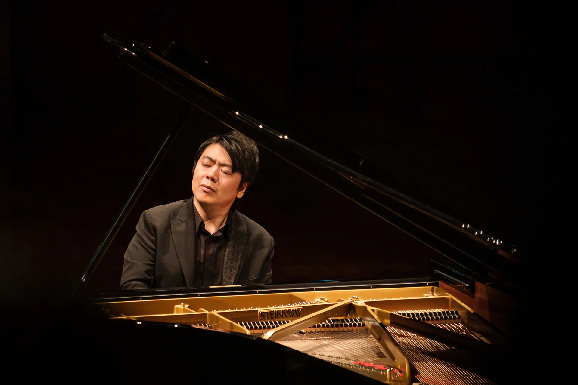 Lang Lang at the grand piano, lit by warm light in the dark hall.