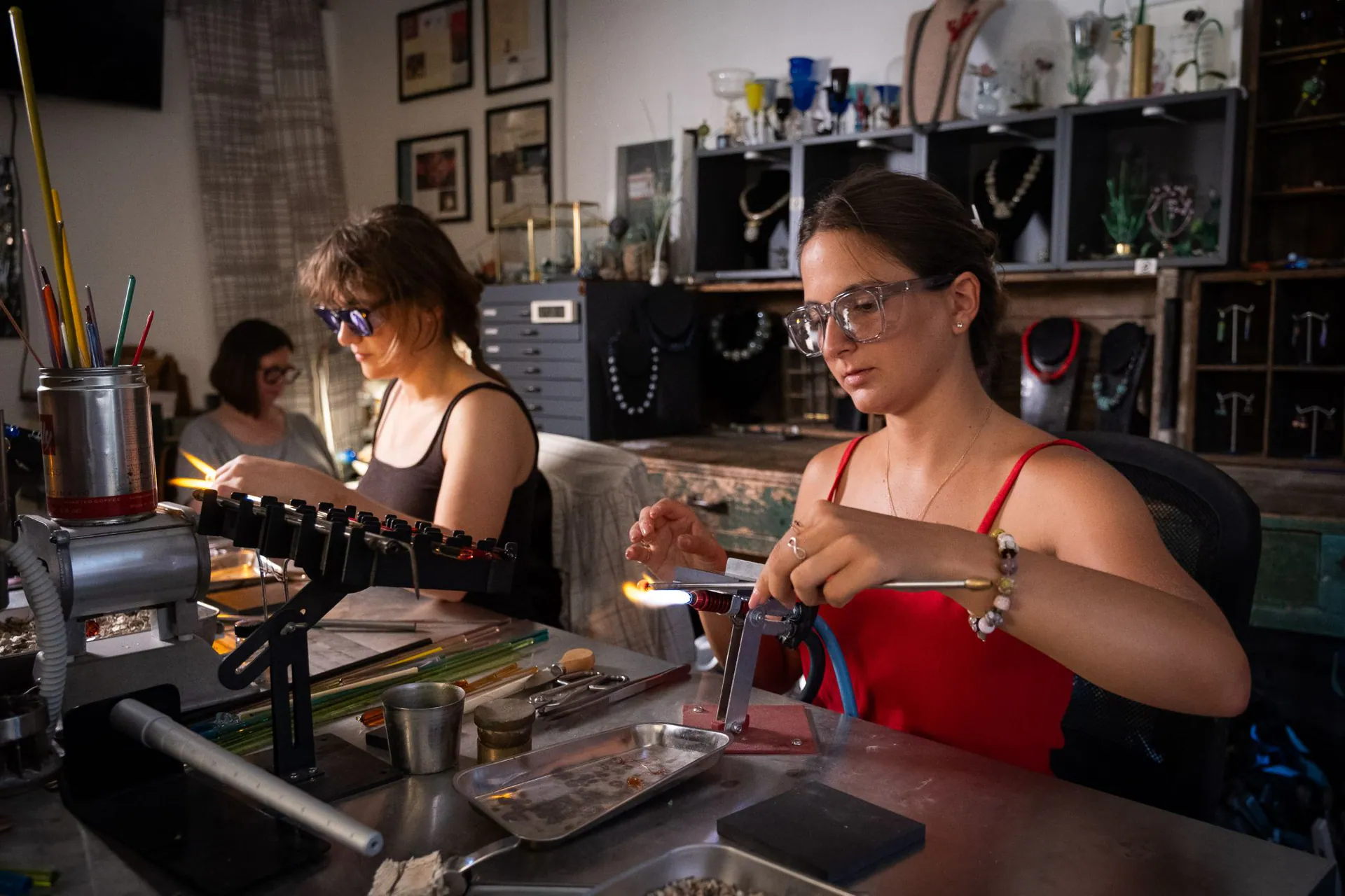 Two young artisans at the lampworking bench, among coloured rods and displayed necklaces