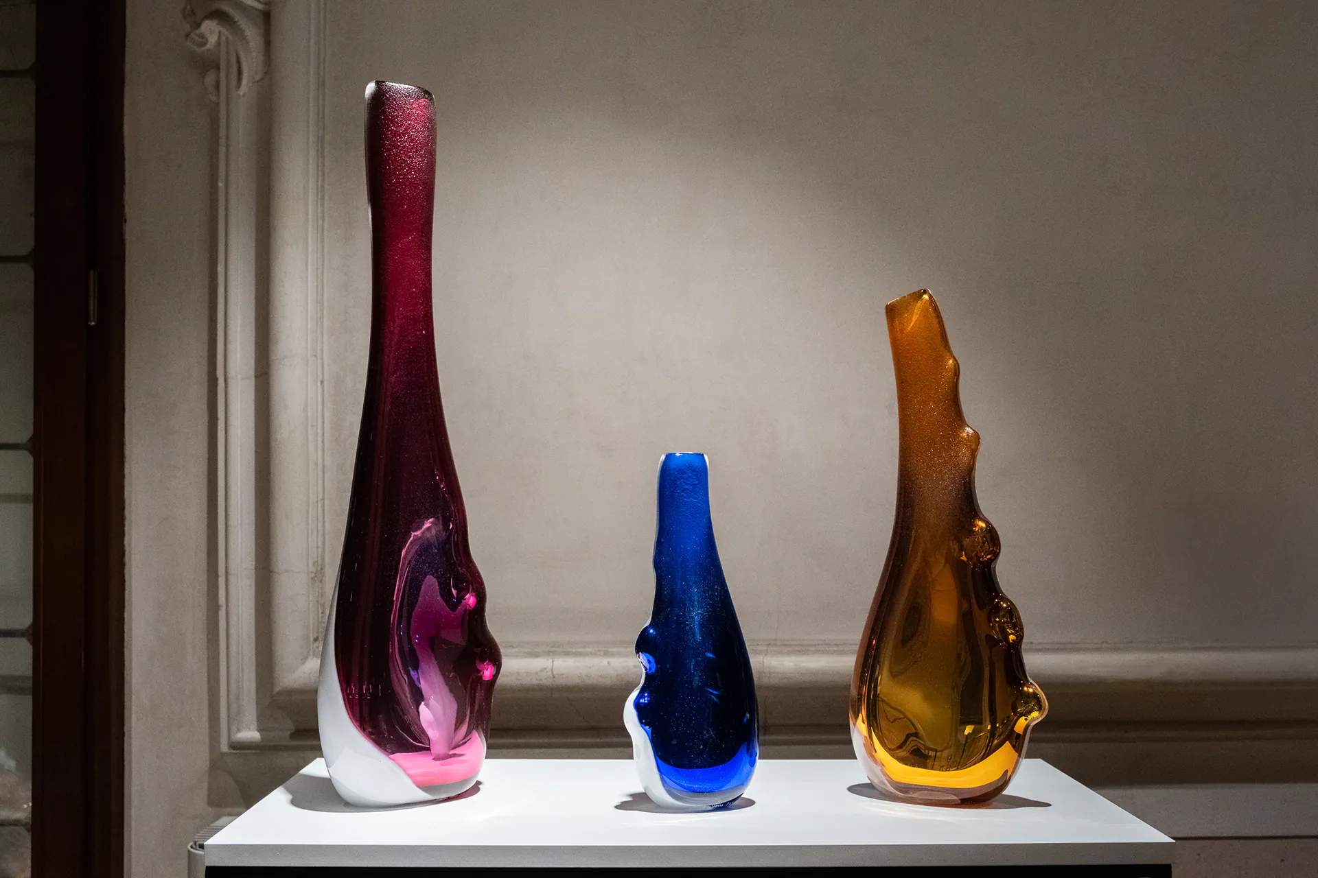 Tre vasi in vetro soffiato, rosso, blu e ambra, esposti su un piano bianco.