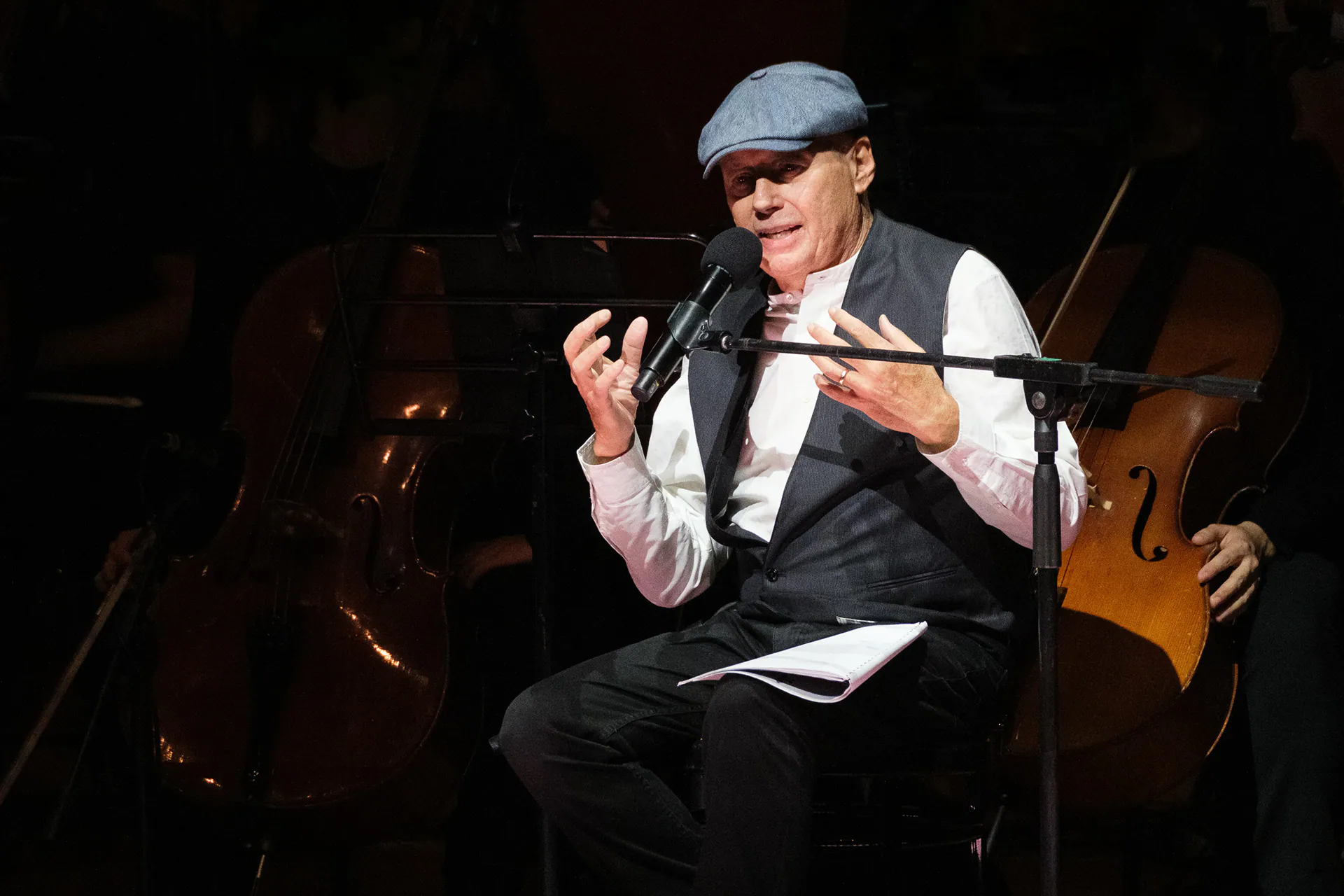 Alessandro Baricco, seated on stage in a cap, narrating into the microphone.