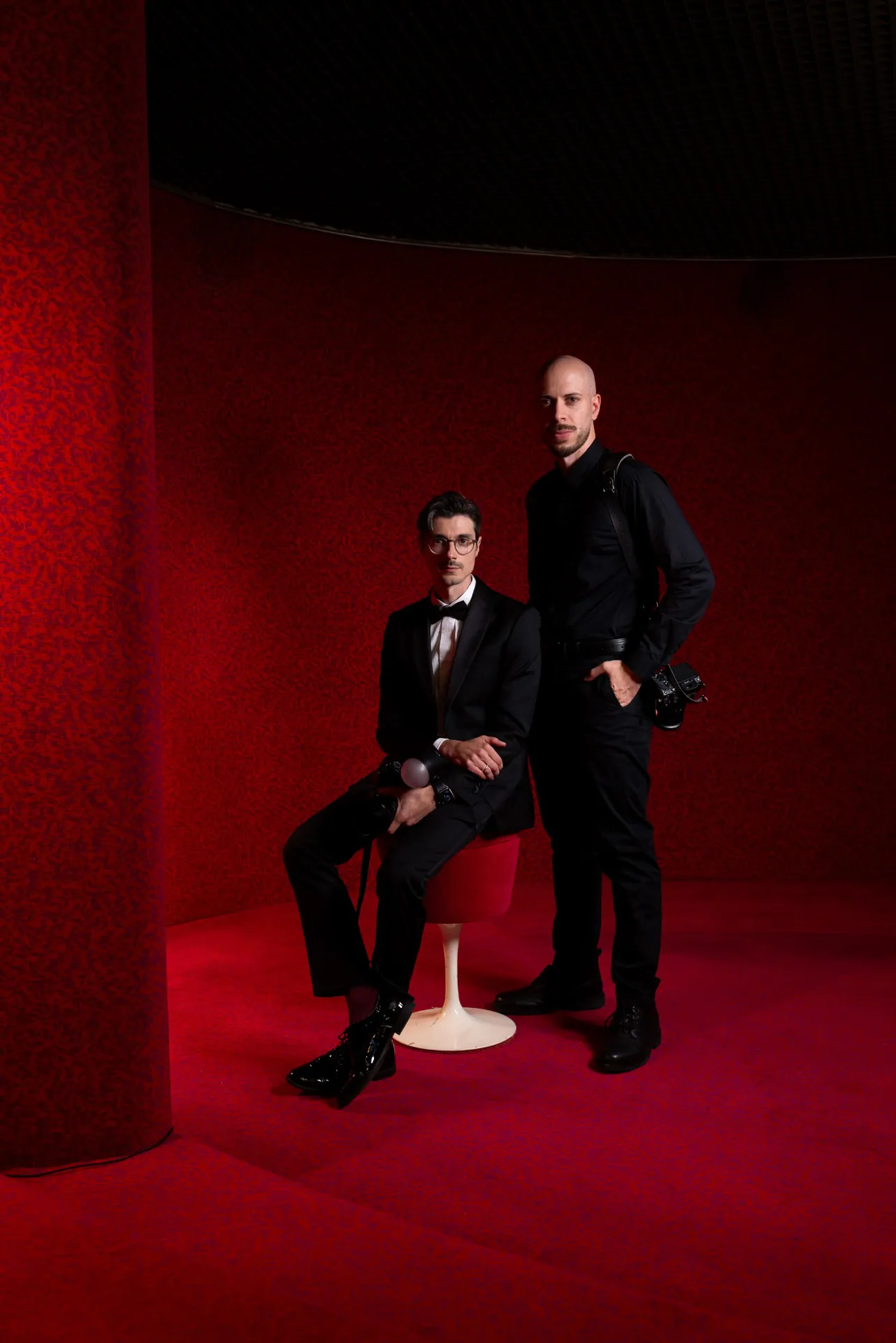Mattia Gaido, seated in a tuxedo, and Simone Borrasi, standing with his camera, in a room lined with red velvet